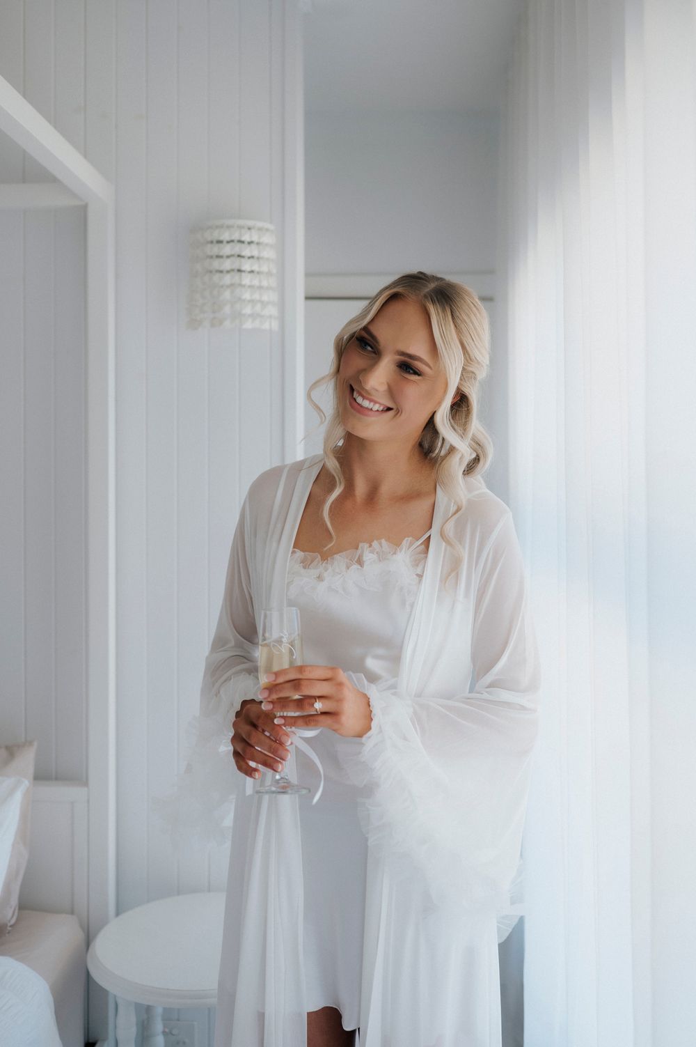 A bride in a white silk robe holds a champagne glass while getting ready on her wedding morning.