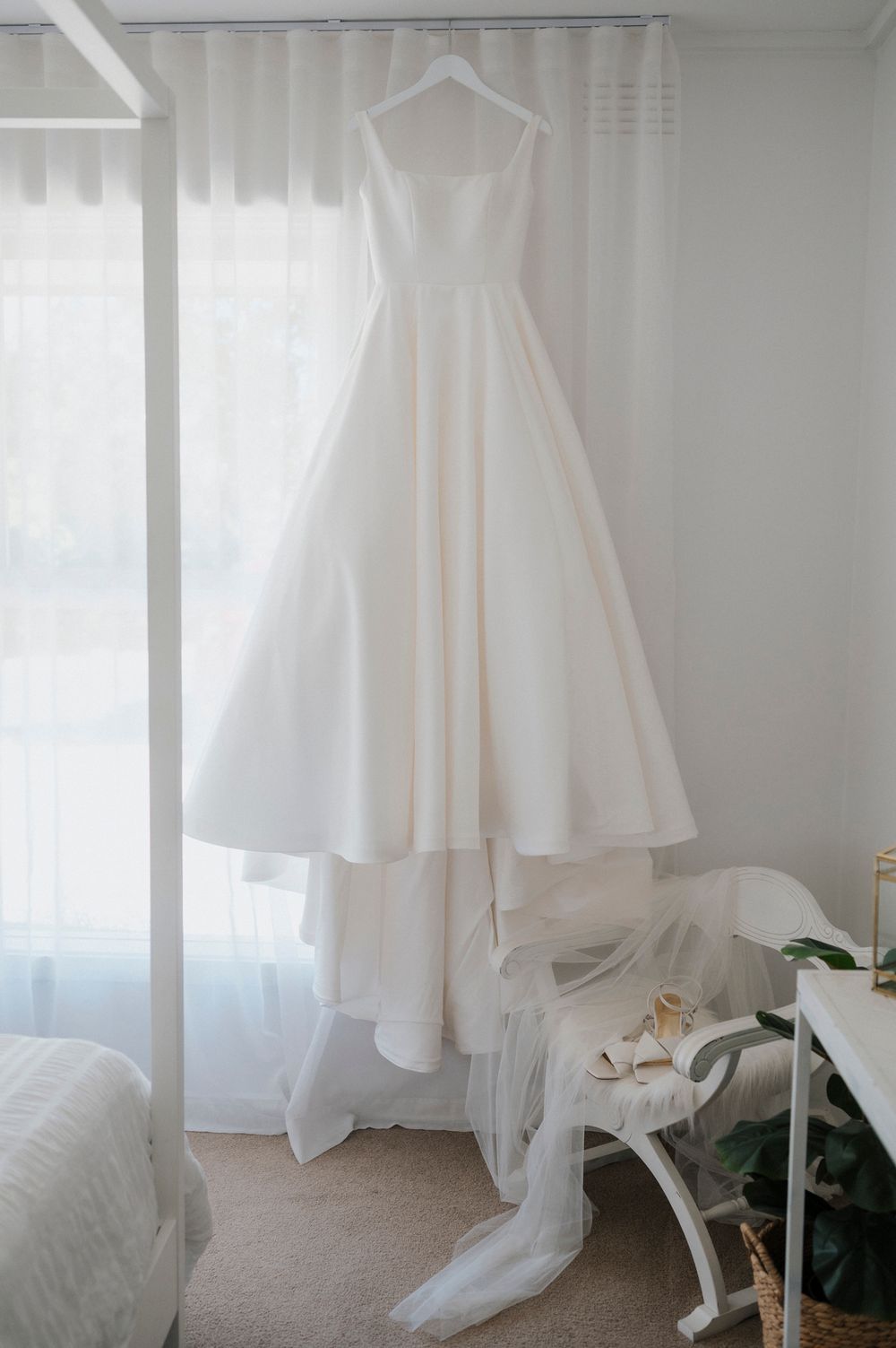 A classic white wedding dress hangs in a bright room with sunlight streaming through sheer curtains.