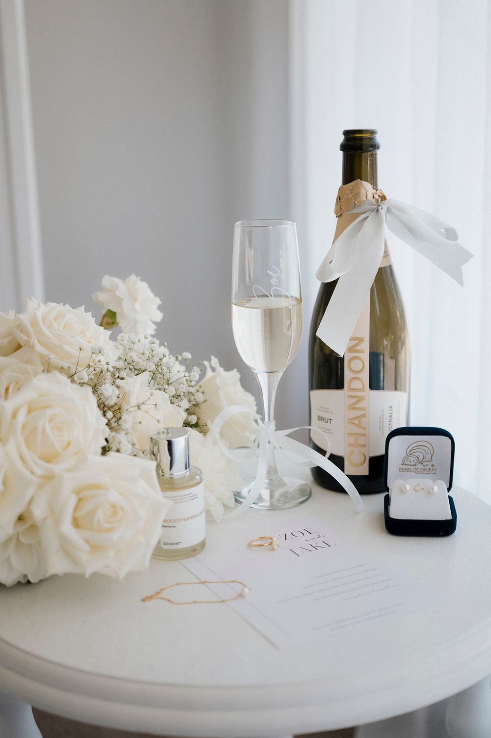 Wedding details including champagne, white roses, and a ring box arranged on a round table.