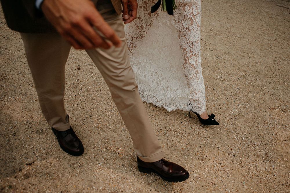 Close-up of groom in khaki trousers and brown leather shoes walking beside bride in lace wedding dress.