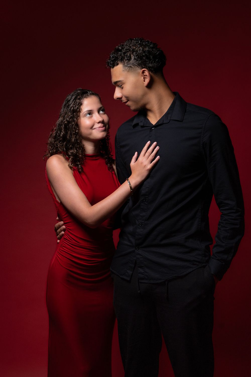 Romantic couple in formal attire embrace against dark red backdrop during elegant photoshoot.