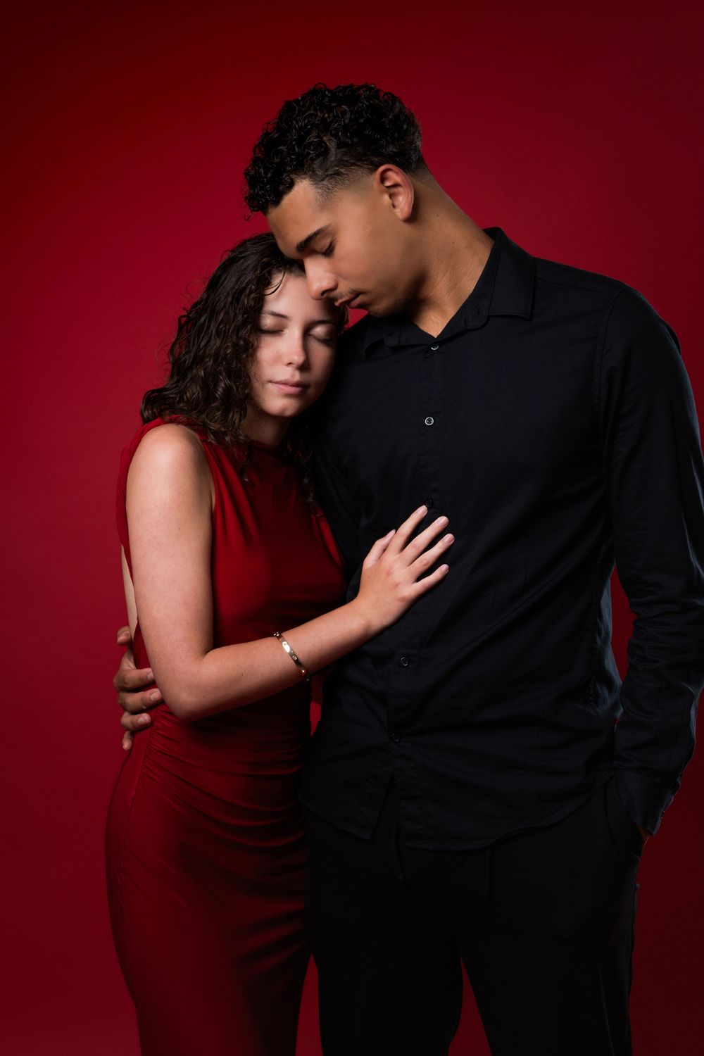 A couple in burgundy and black poses intimately against a dramatic dark red background for romantic portraits.