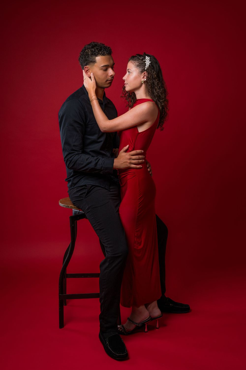 Romantic couple in black and red attire pose intimately against dark red backdrop with dramatic studio lighting.