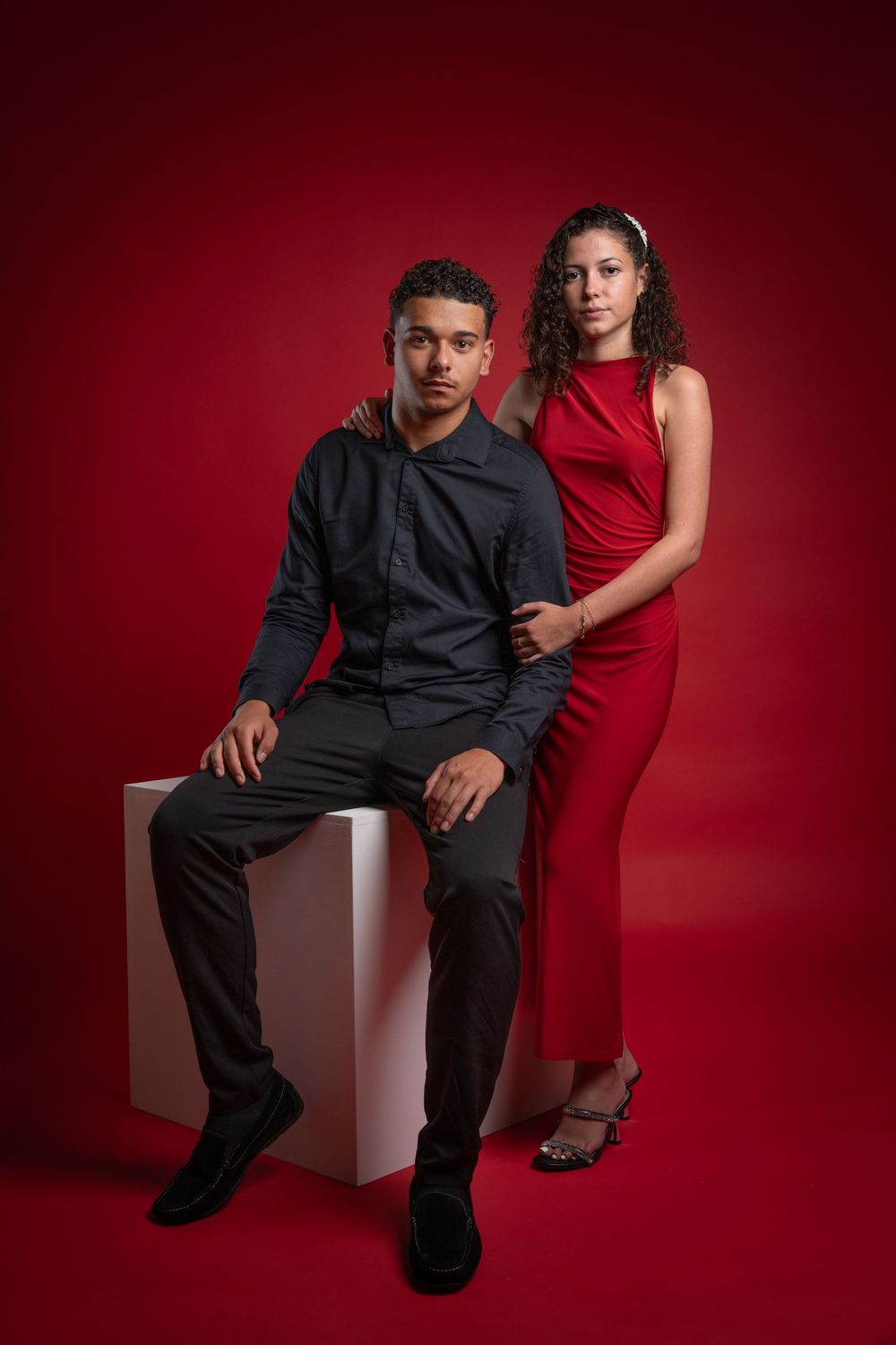 Artistic portrait series against deep red background showing couple in contrasting red dress and black outfit posing on white cube.