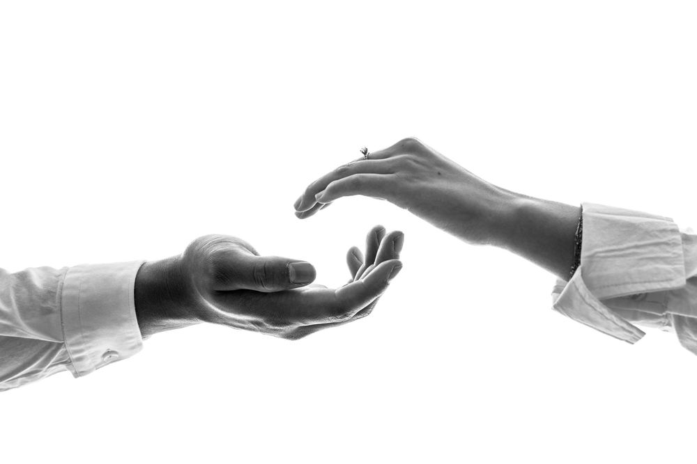 Black and white artistic photograph of two reaching hands in white sleeves against a white background.
