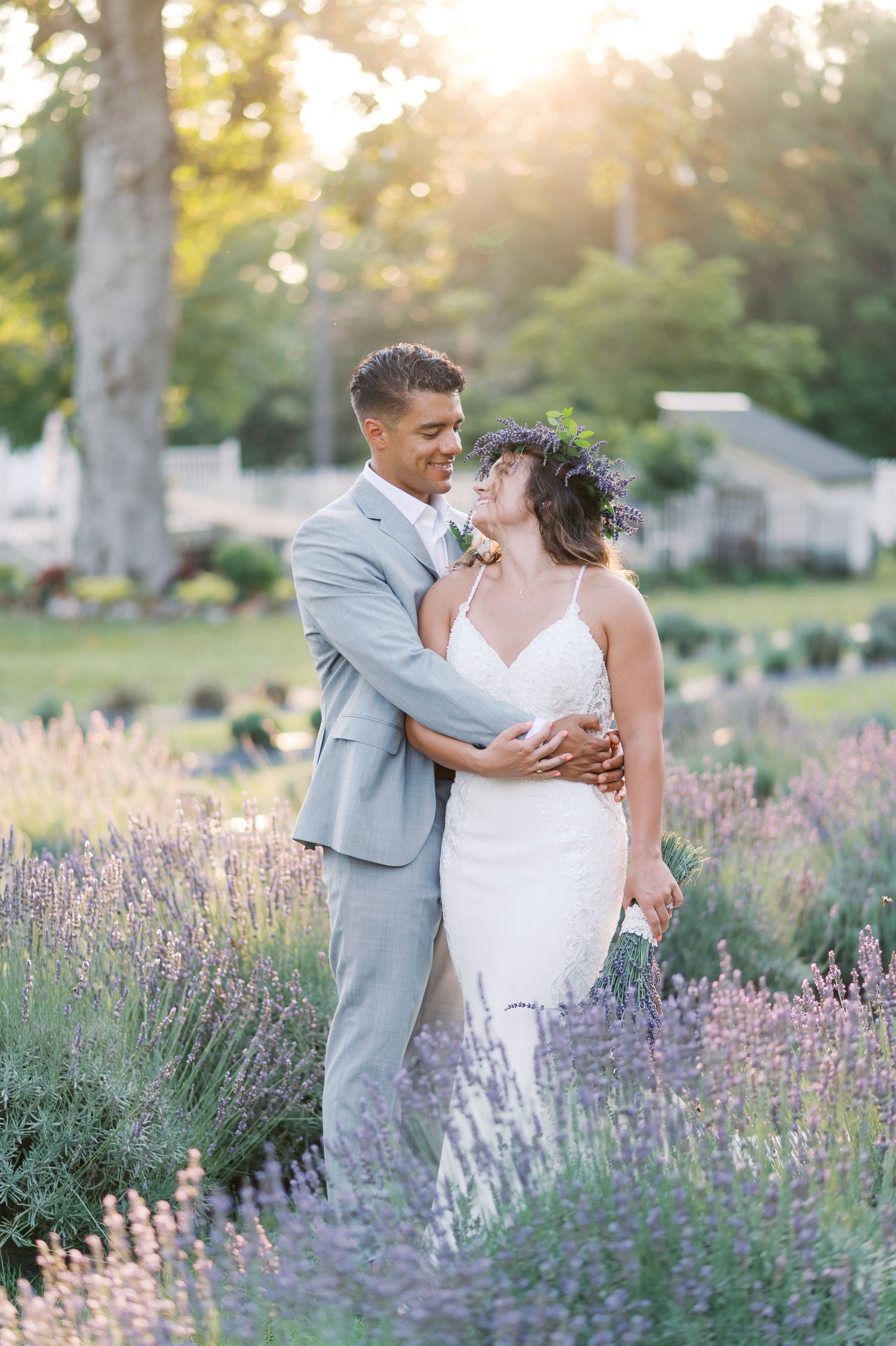 A romantic wedding couple embraces in a blooming lavender field at sunset with soft golden light filtering through trees.