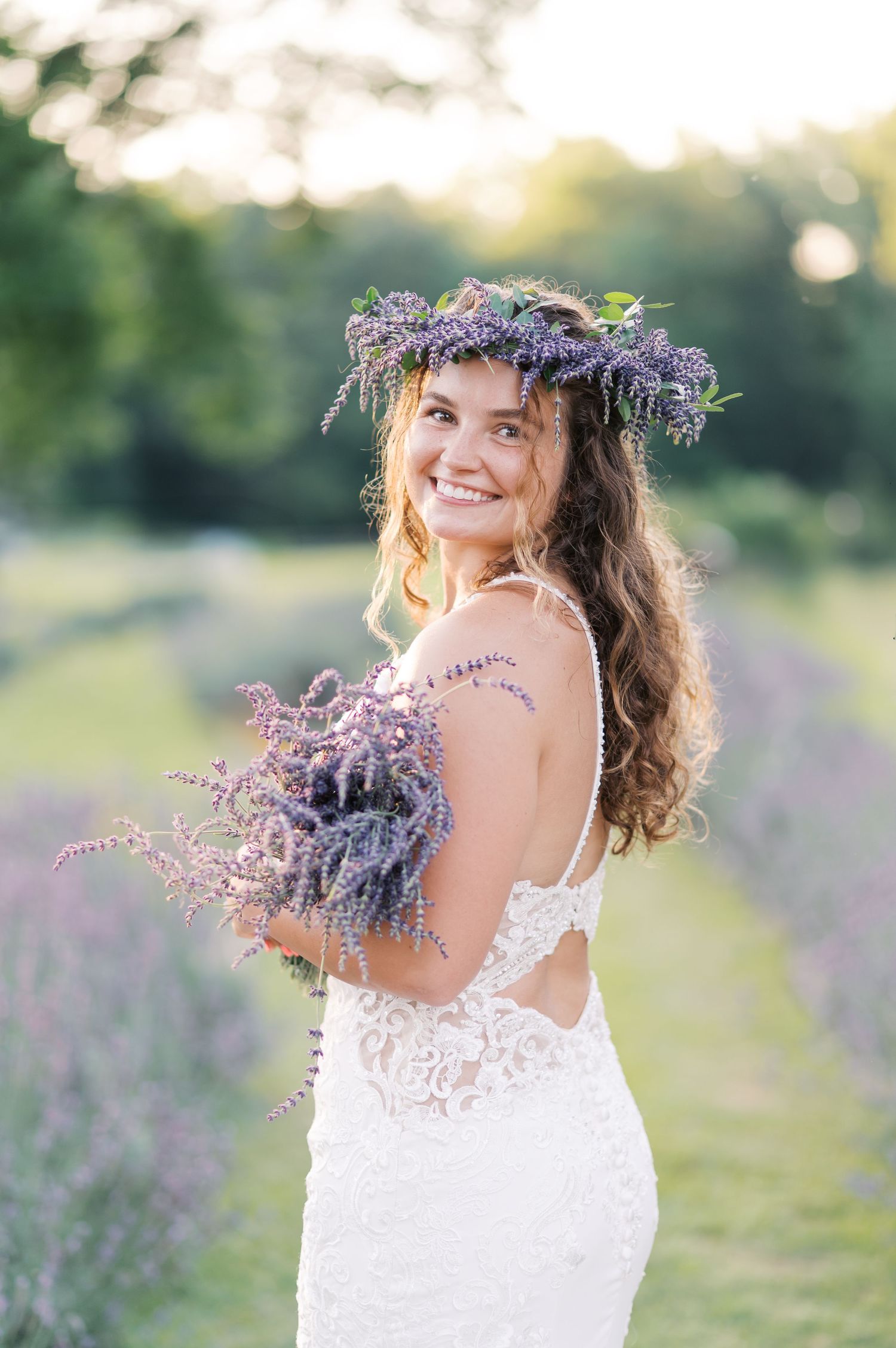 A bride wearing a white lace dress holds a bouquet of purple lavender flowers in a lavender field at sunset.