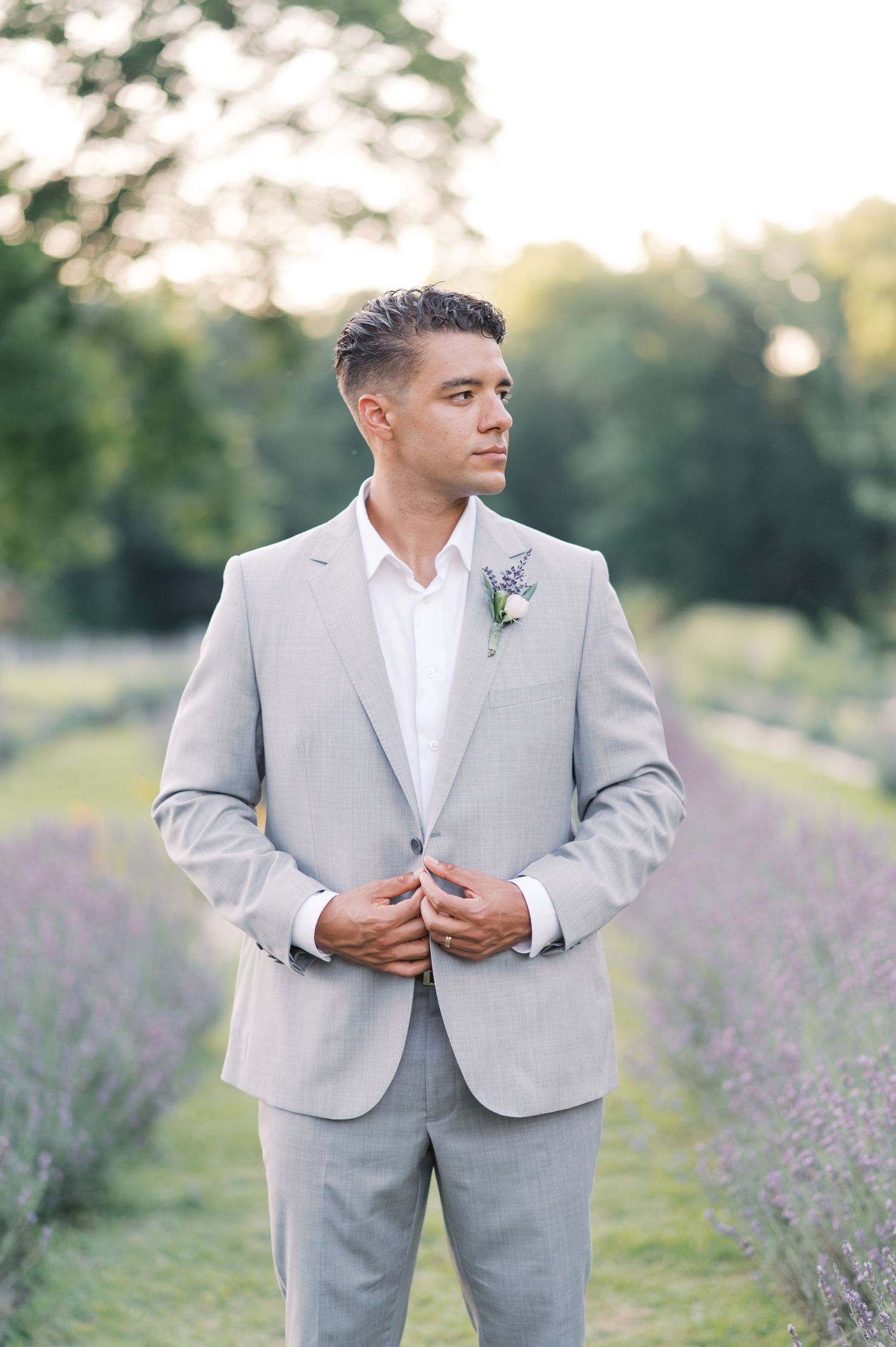 A groom wearing a light gray suit and white boutonniere stands in a dreamy lavender field at golden hour.