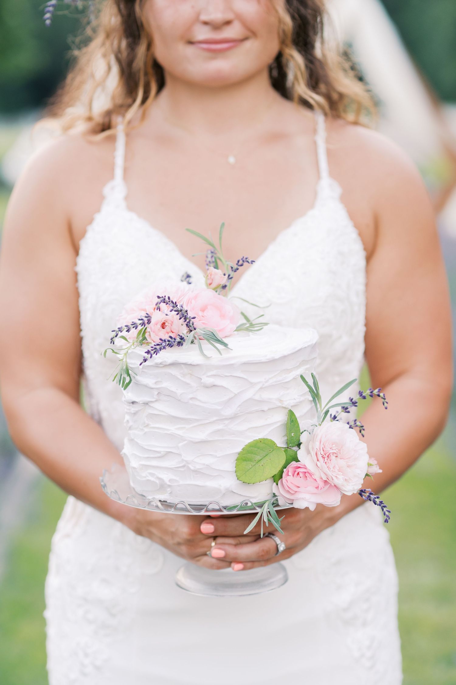 Bride holding a simple but beautiful wedding cake adorned with light pink roses and sprigs of lavender,