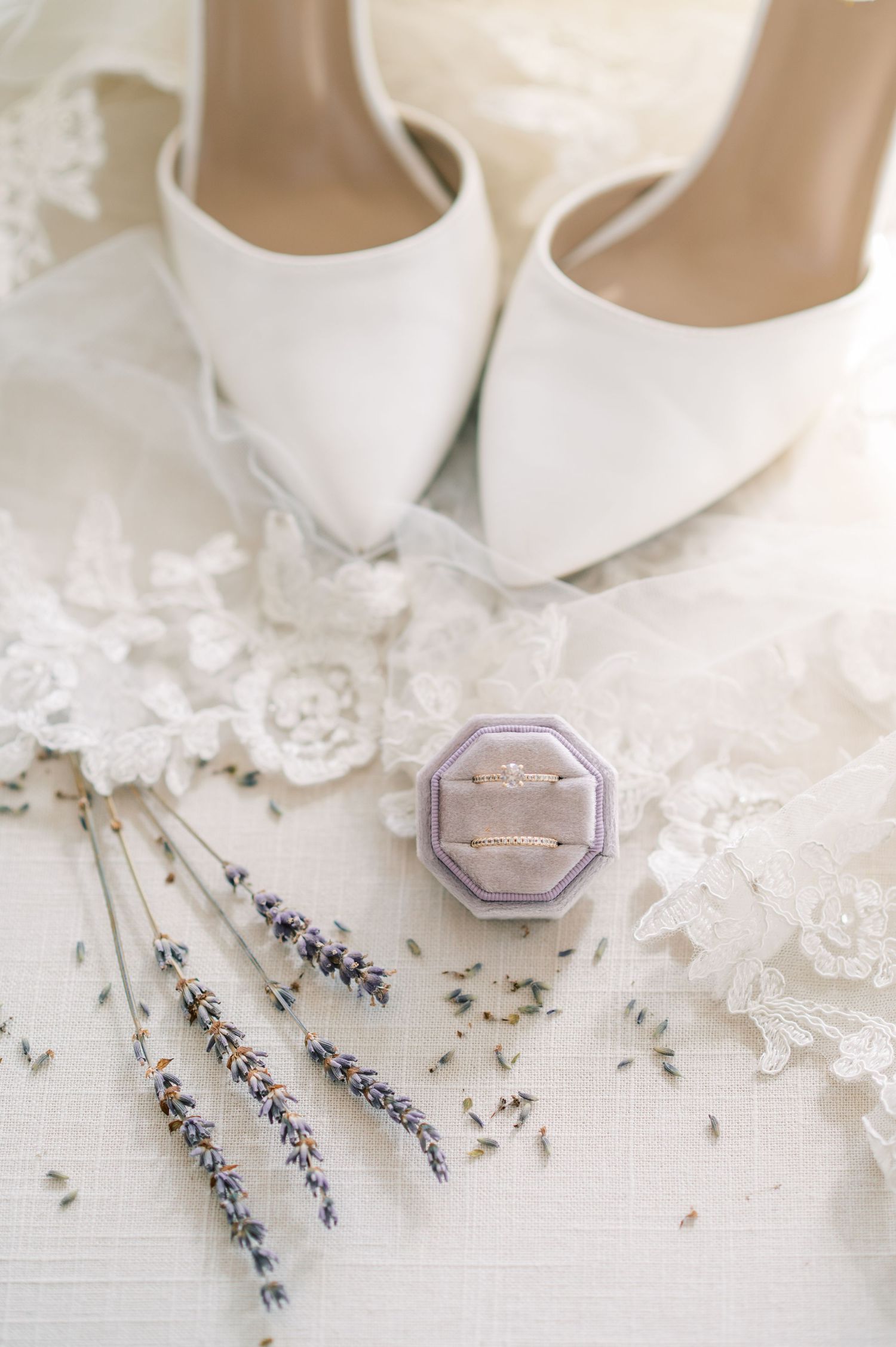 Elegant white bridal shoes arranged with lavender sprigs and jewelry on delicate lace background.