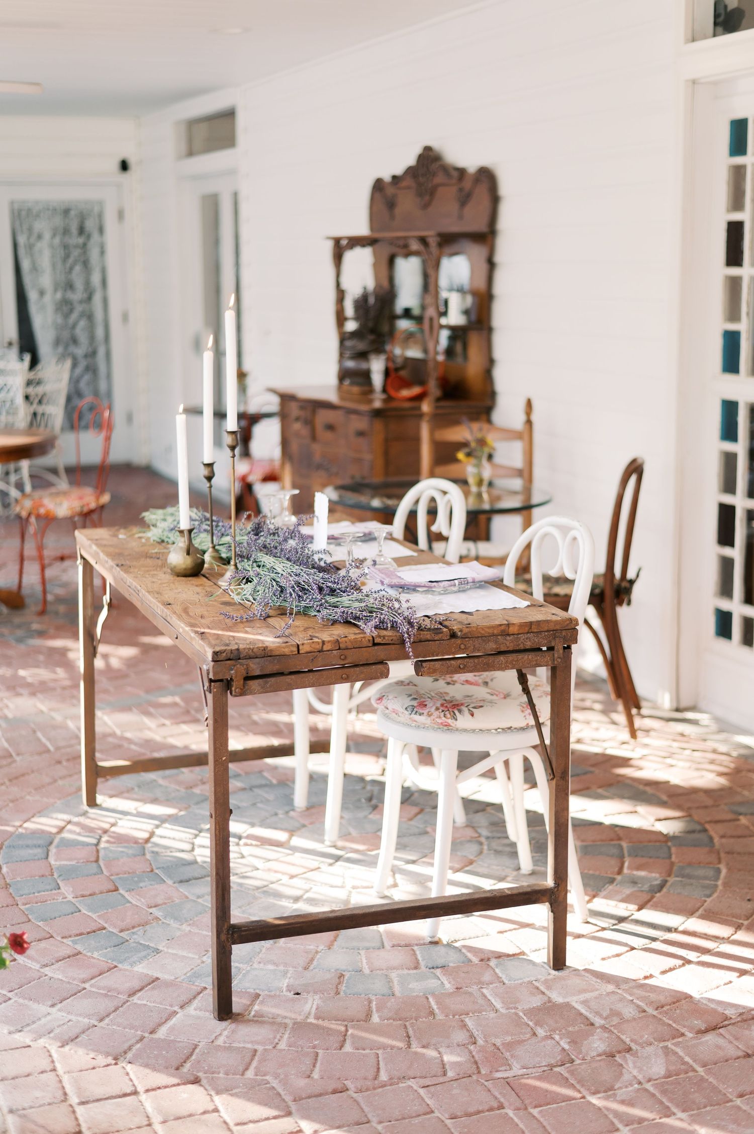 Rustic wooden dining table with white chairs on a brick patio showcasing vintage decor and ornate mirror in background.