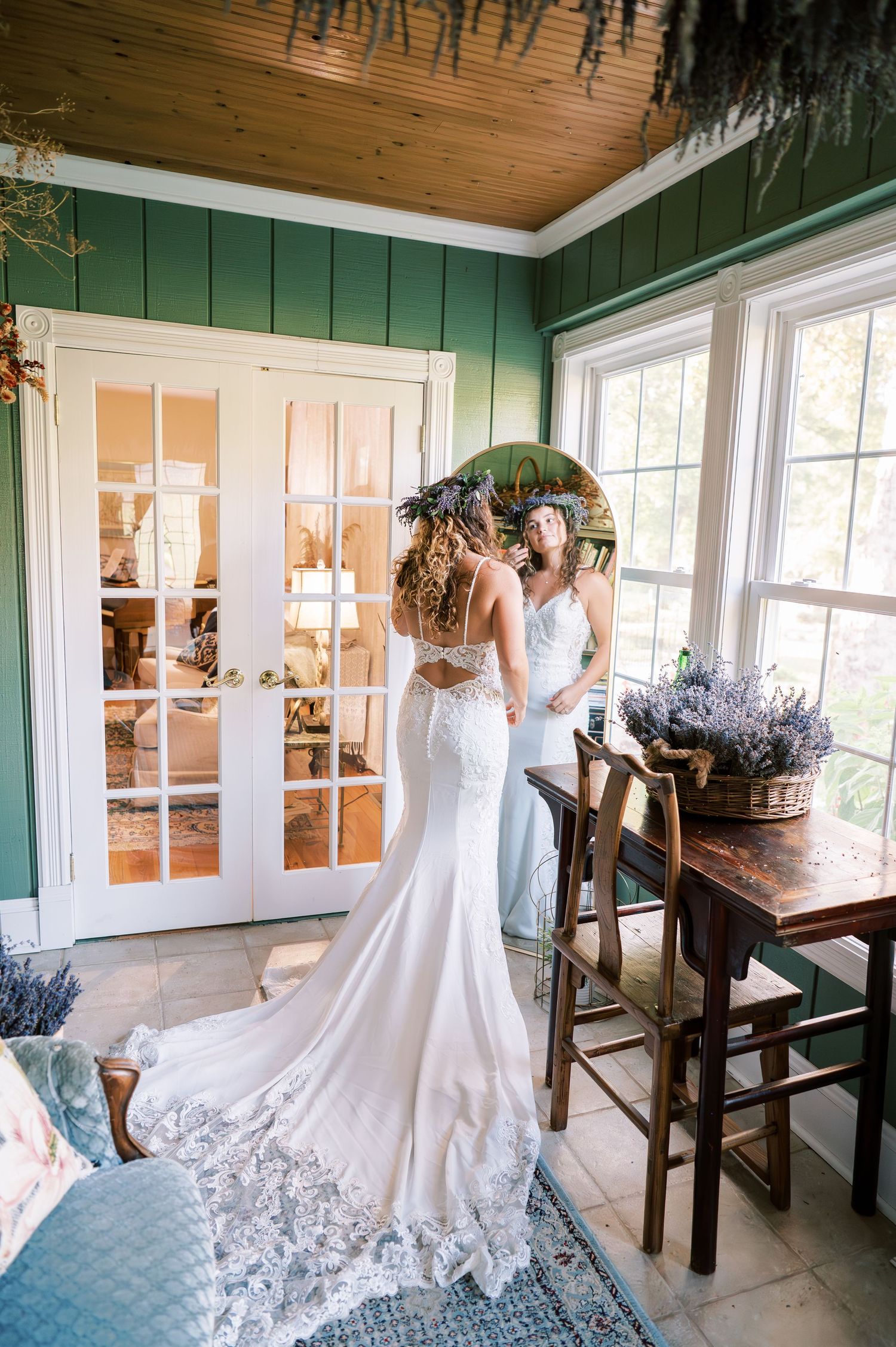 White wedding dress with intricate lace details shown in natural light inside a rustic room with green walls and wooden floors.