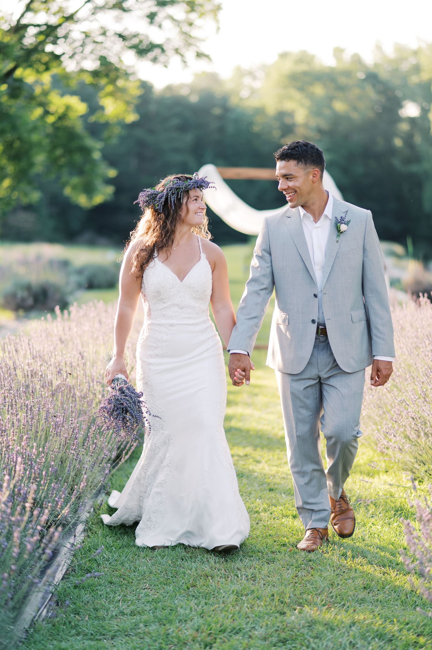 Romantic newlyweds hold hands while walking through a lavender field at sunset in elegant wedding attire, near Raleigh NC