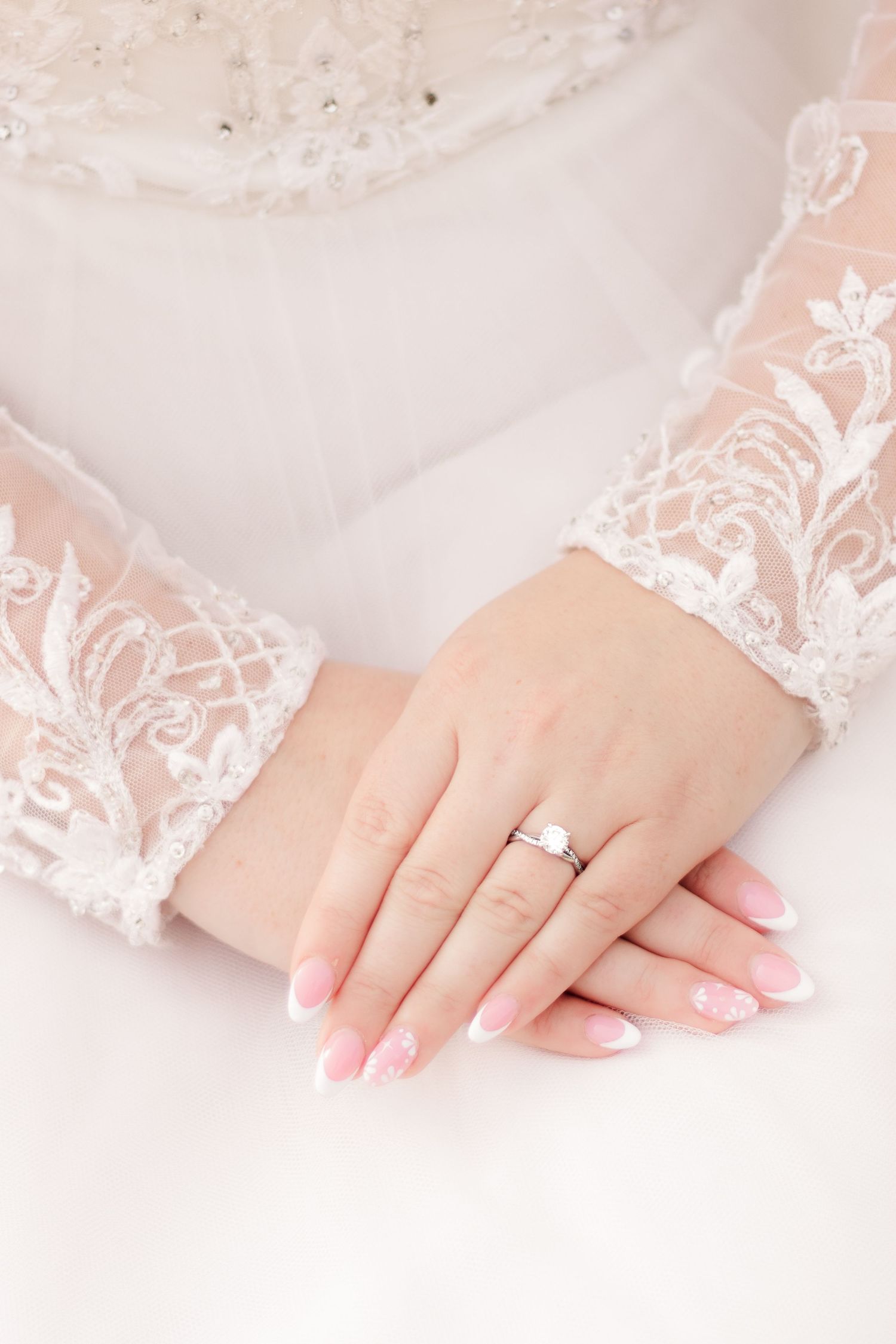 Close-up of delicate lace sleeves and engagement ring on hands against white background.