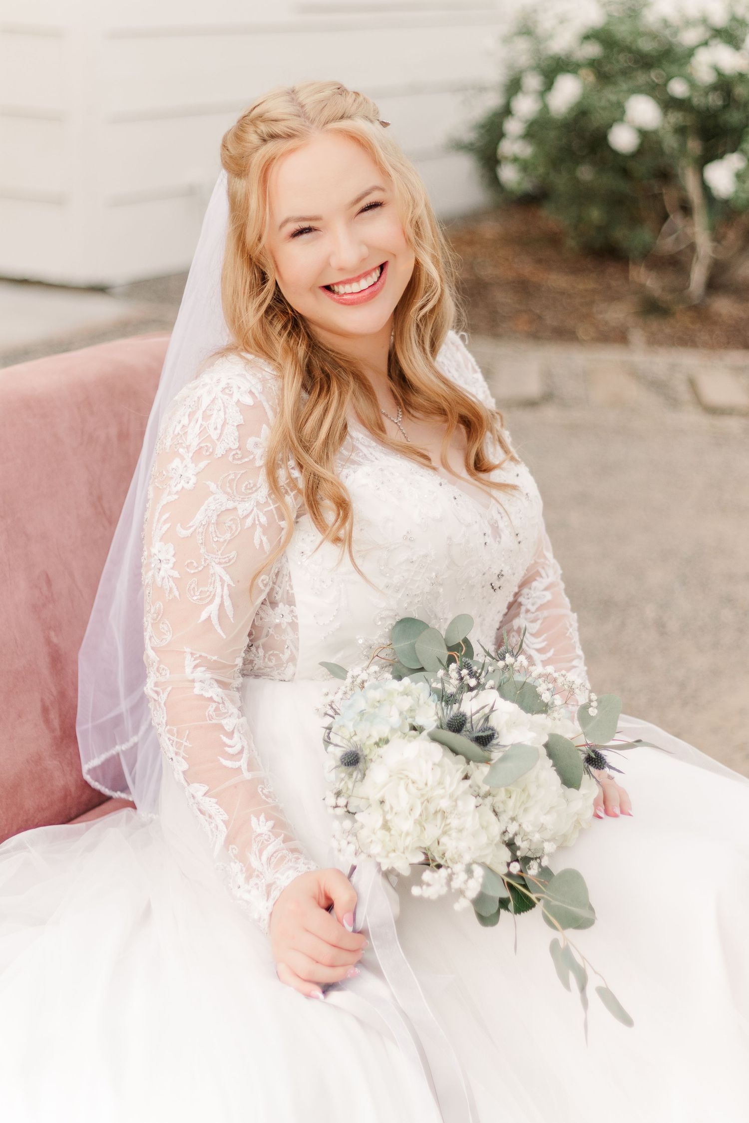 A bride in a long-sleeved lace wedding dress sits on a pink chair holding a white and green bouquet.