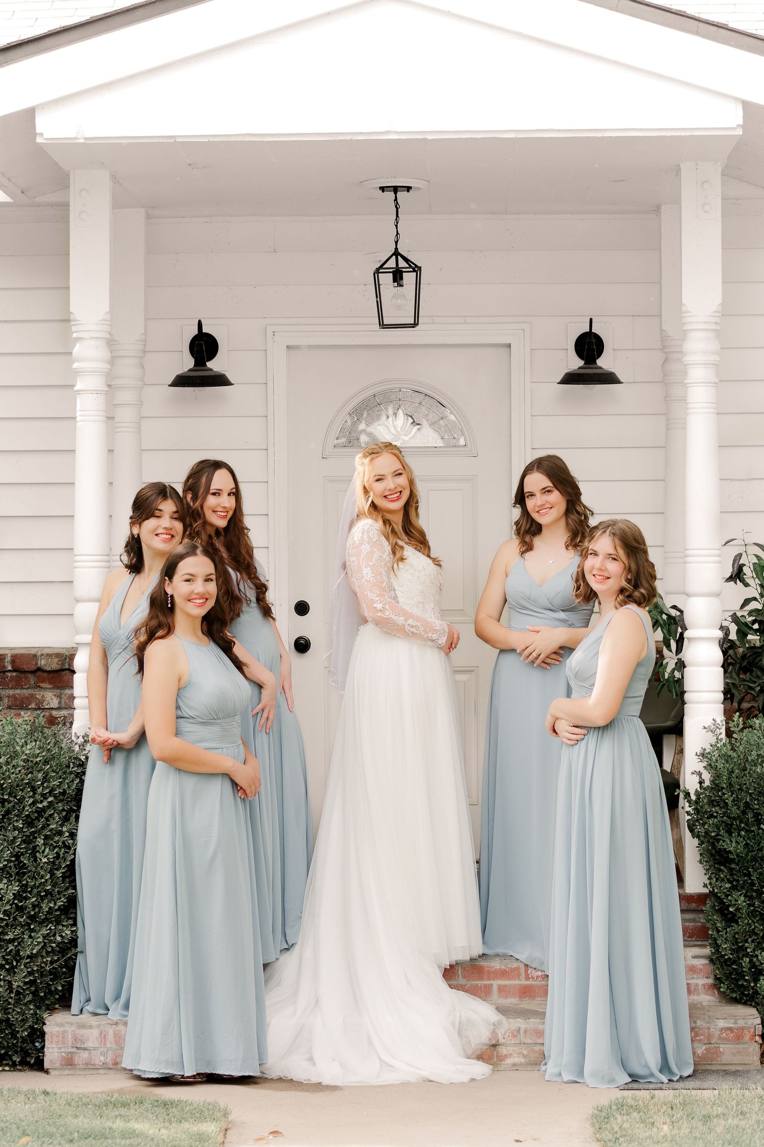 A bridal party poses on a white porch with bridesmaids wearing light blue dresses.