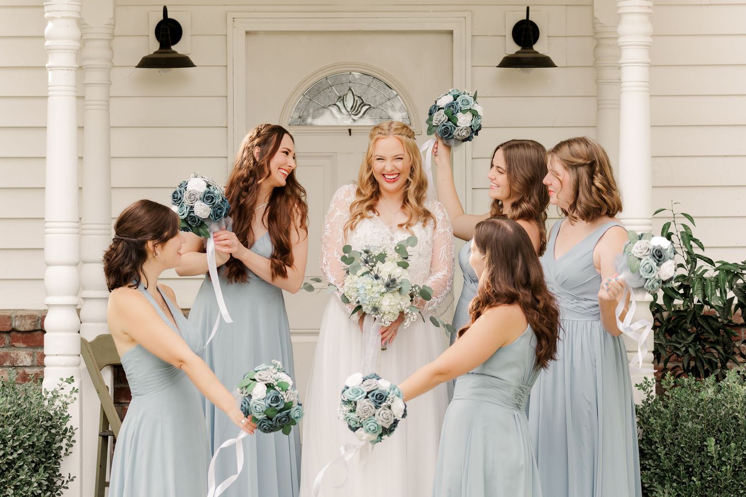 Bridesmaids in soft blue dresses hold bouquets while posing outside a white building.