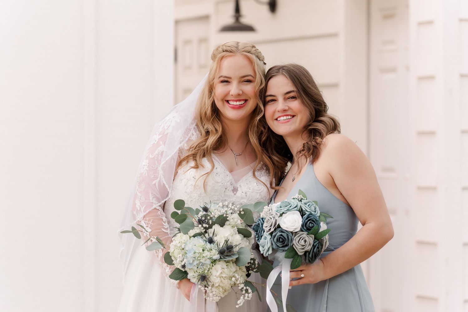 Wedding party members in light blue dresses pose with bouquets against white building exterior.