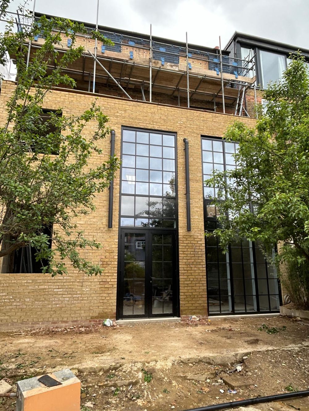 Modern building renovation showing tall glass Crittall style windows and exposed brick exterior with scaffolding on the top floor.