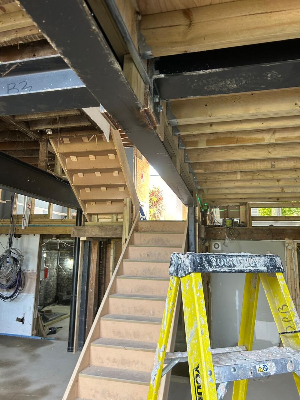 Interior basement renovation showing wooden stairs, exposed beams, and a yellow ladder in an unfinished space.