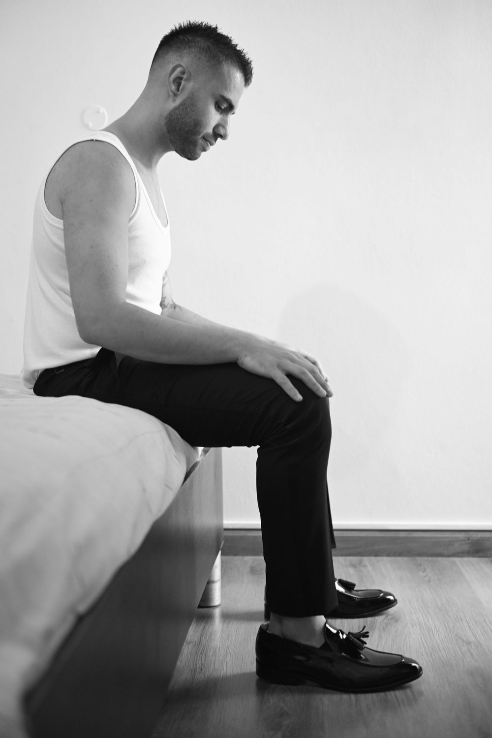 Person in white tank top and black pants sitting on edge of bed looking down.
