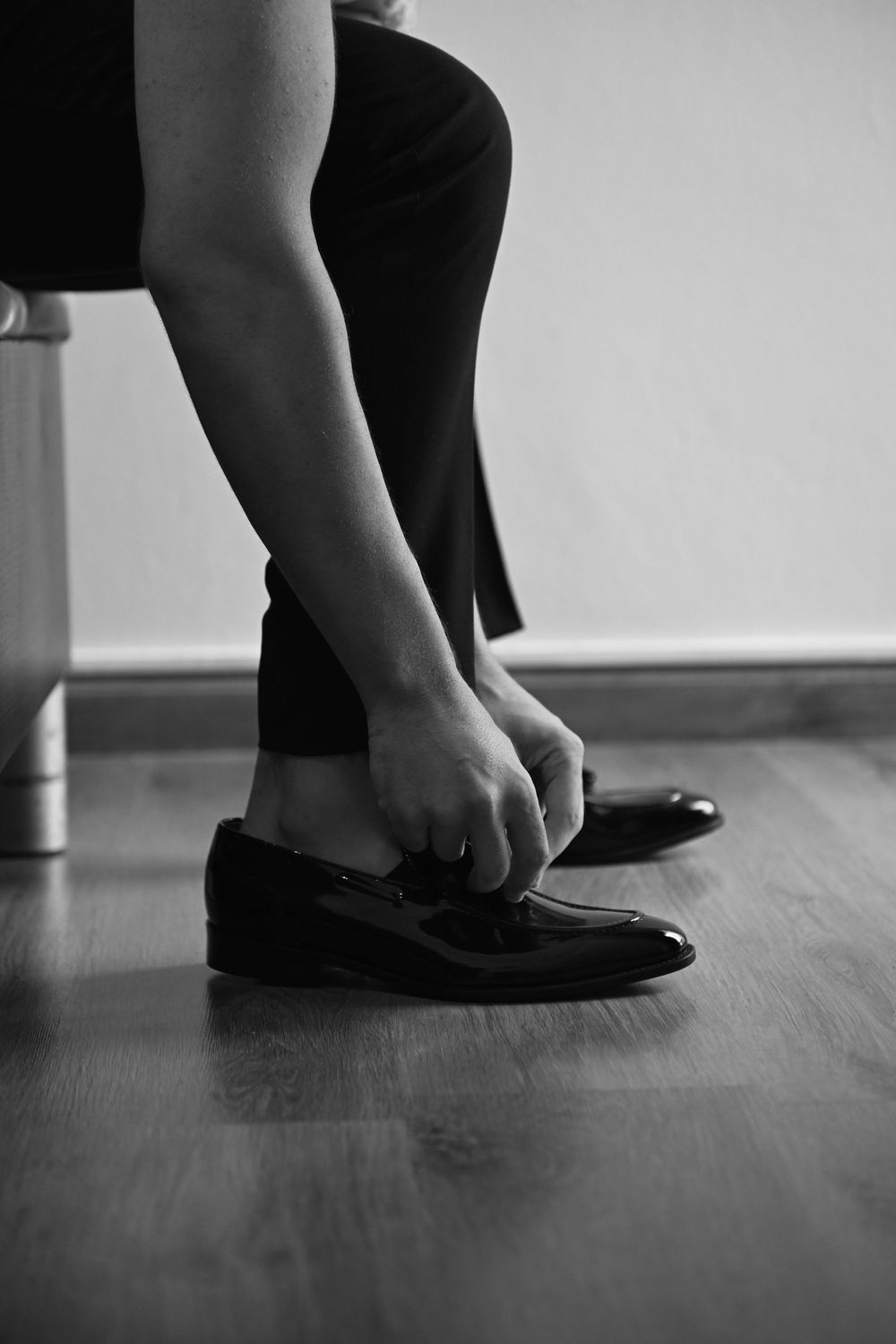 Close-up of hands adjusting black leather loafers on wooden floor.