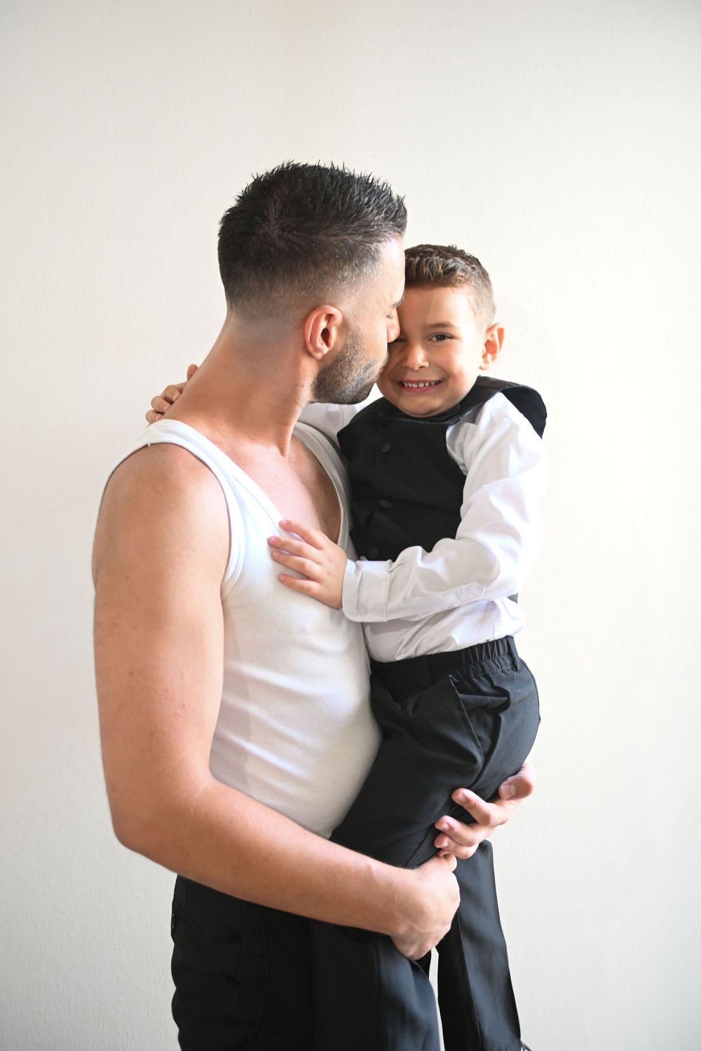 Tender moment between parent and child in formal wear against white wall.