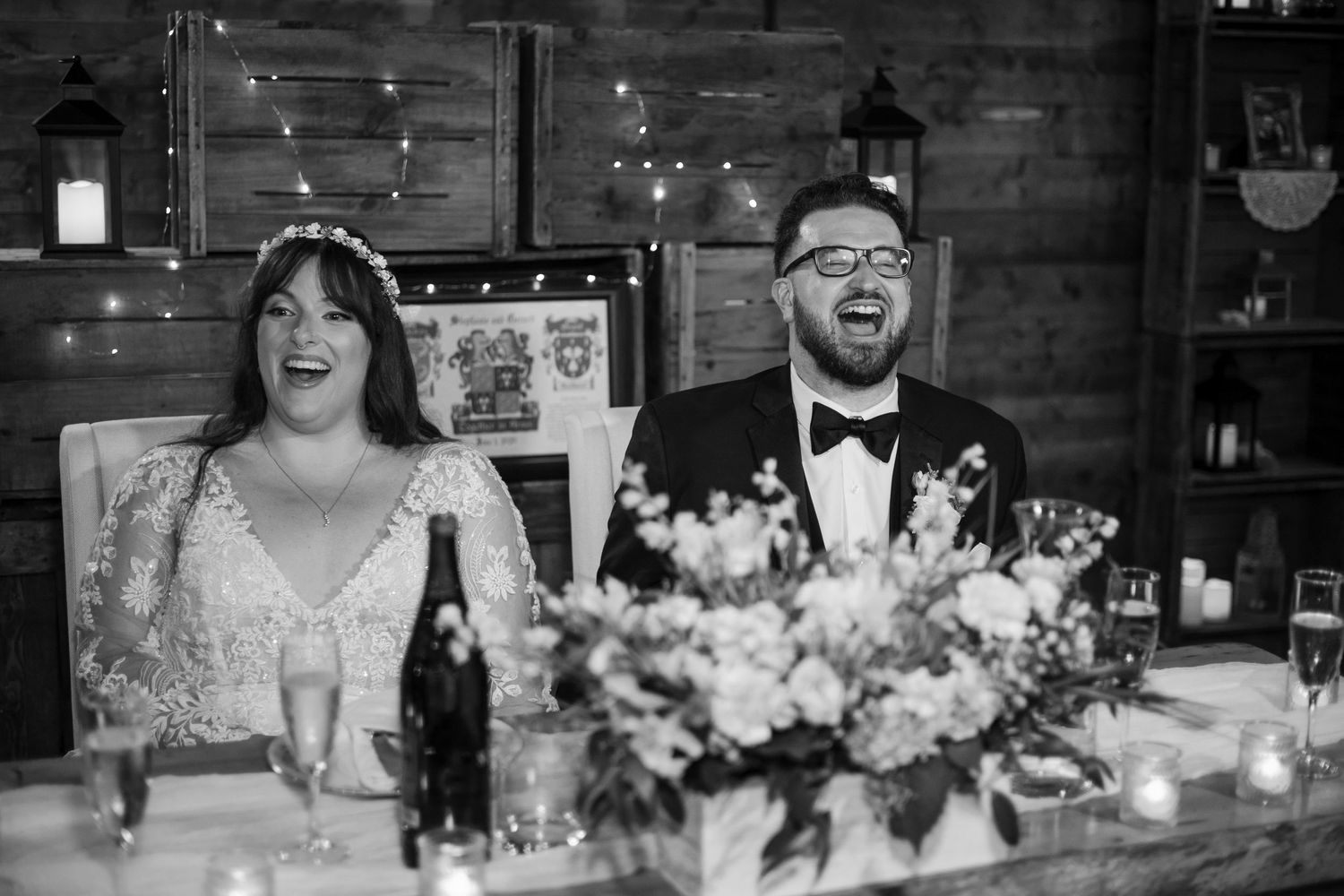 Wedding guests seated at long rustic tables illuminated by string lights in a barn reception venue.