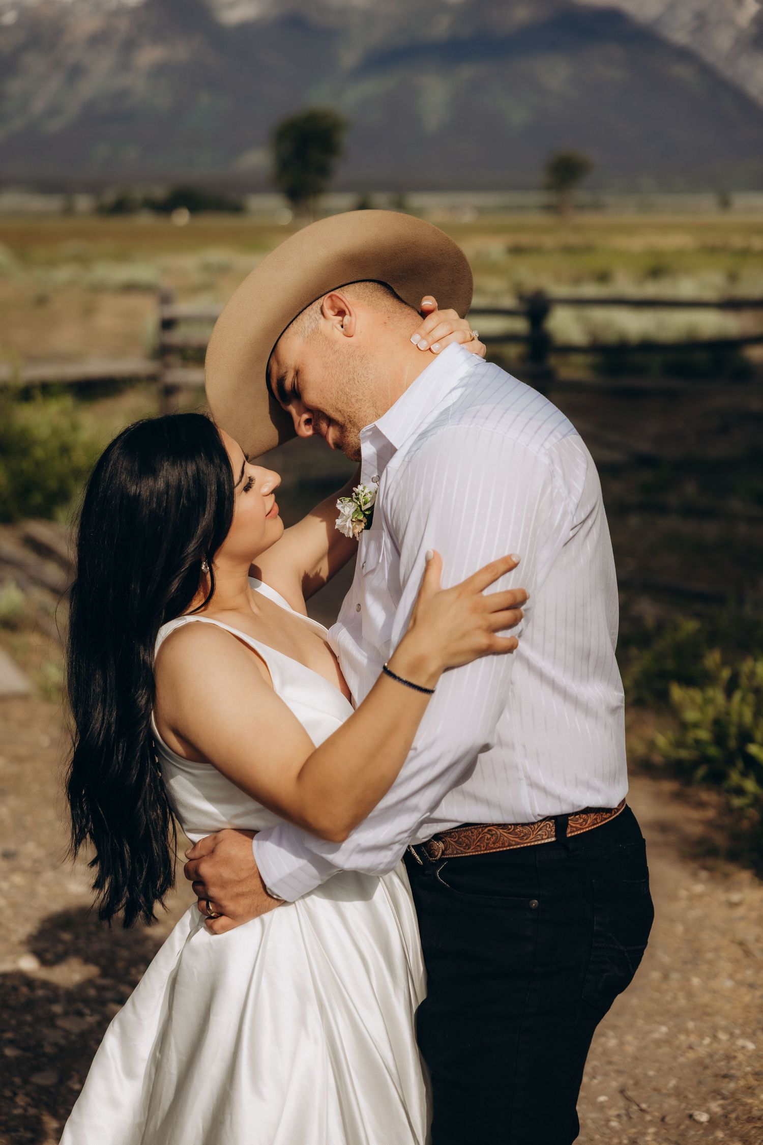 A romantic couple in western wedding attire embrace against a mountain landscape background in golden sunlight.