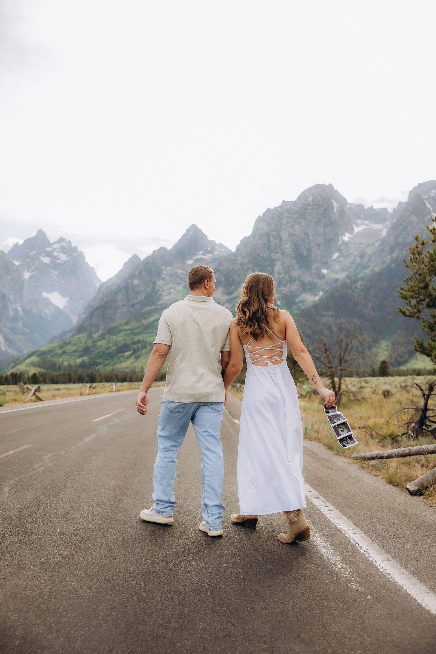 Couple walking on mountain road with scenic Grand Teton peaks in background wearing casual summer attire.