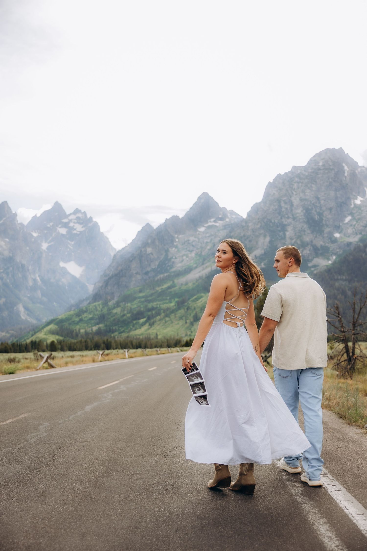 A couple walks on a mountain road with the majestic Grand Teton peaks rising in the background.