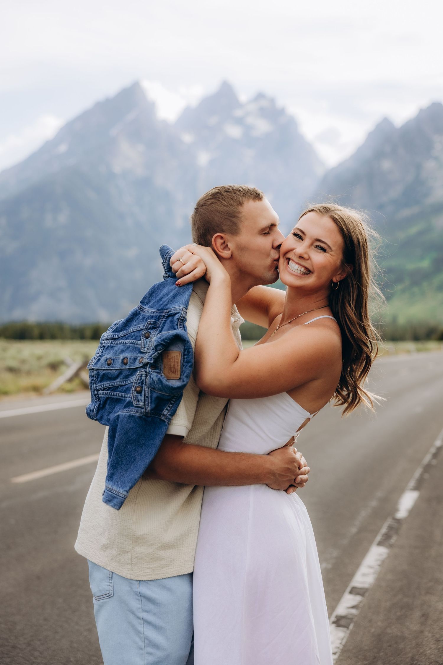 A couple embraces on an empty road with majestic mountain peaks in the background.