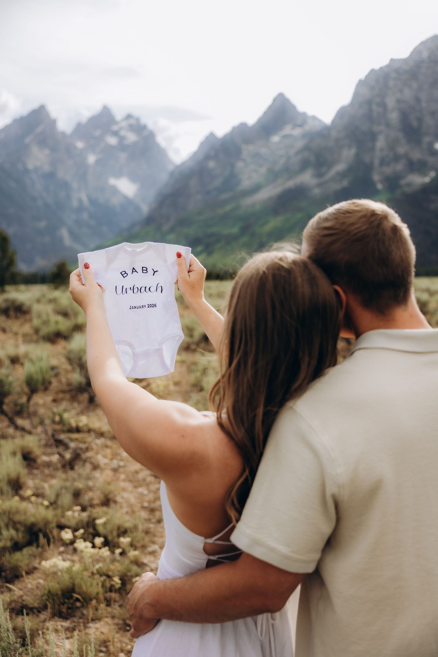A couple holds up a baby onesie against a backdrop of majestic mountain peaks in Grand Teton National Park.