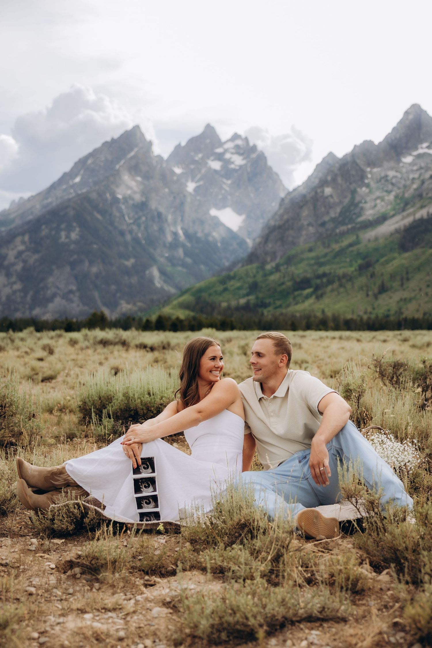 A couple relaxes in a wildflower meadow with dramatic Teton mountain views behind them.