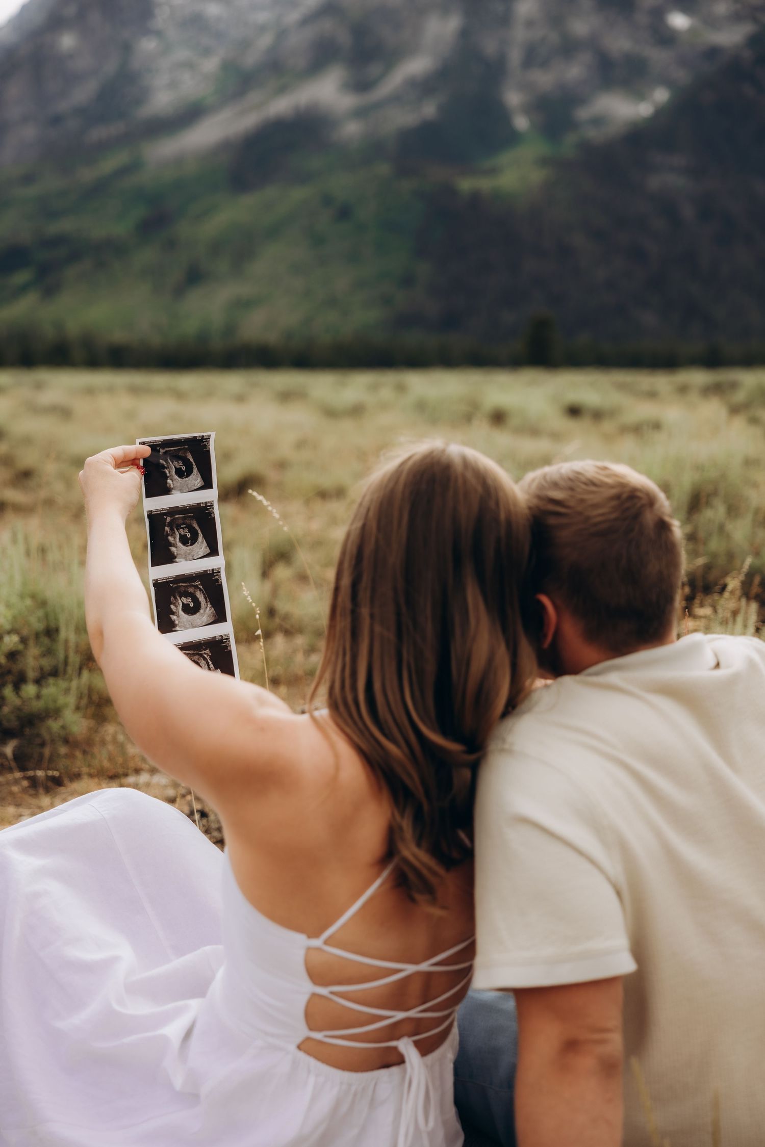 Two people holding up an ultrasound photo against a backdrop of mountains and open fields.
