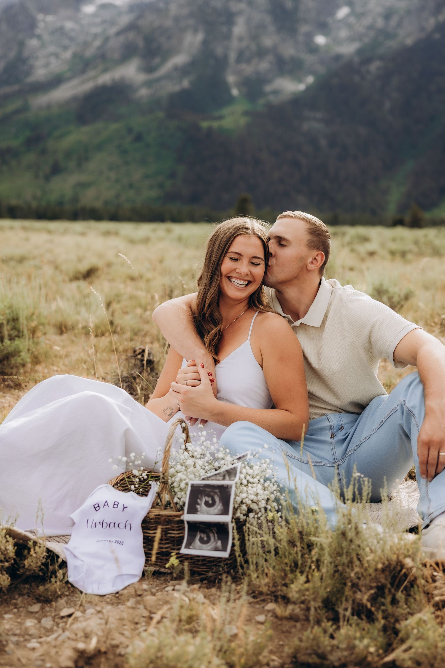 Romantic couple poses together in a mountain meadow with golden light and vintage camera for a pregnancy announcement photoshoot.