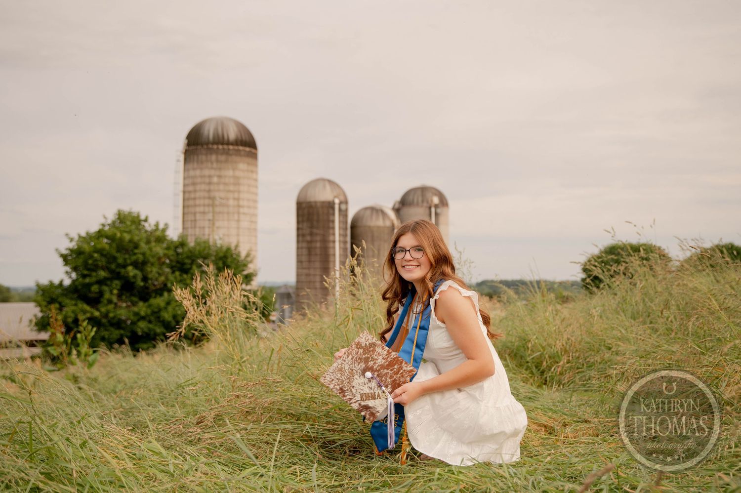 Farm portrait session with a rustic backdrop featuring tall grain silos and green summer grass in a countryside setting.