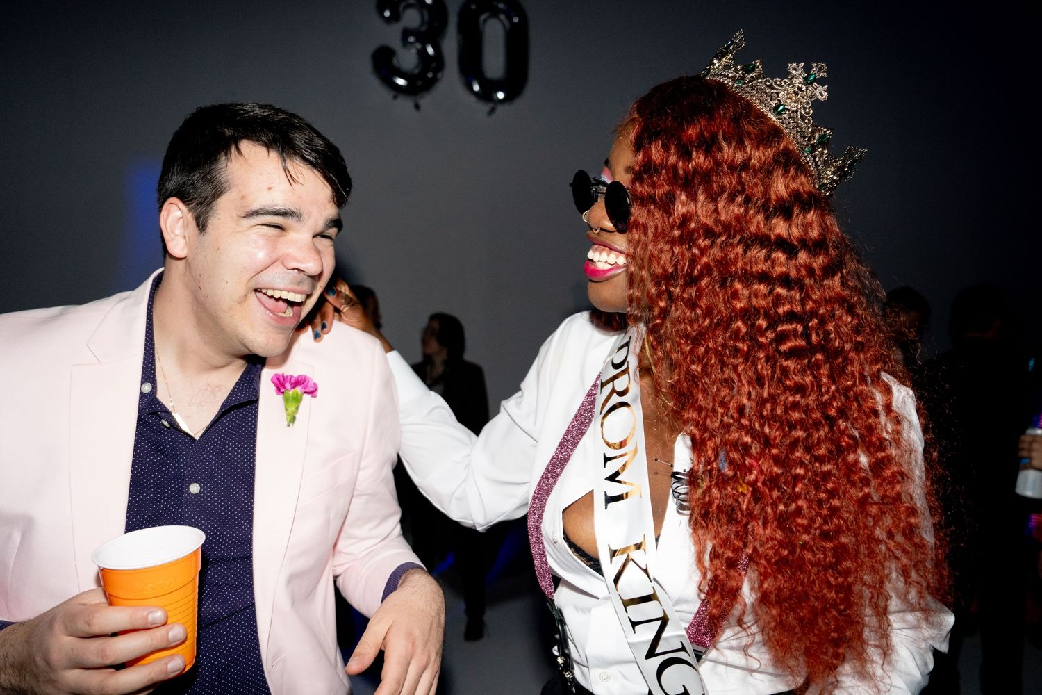 Birthday Prom Bash: Two people stand laughing in a dark club, one is wearing a crown and Prom King sash while laughing at the one in pink