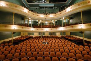 Interior view of a historic theater with multiple balcony levels and rows of orange velvet seats facing an empty stage.