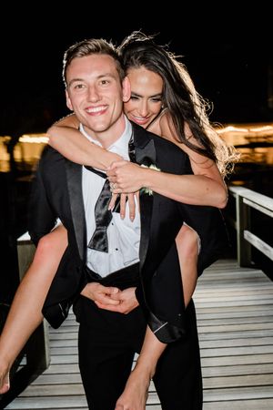 A joyful couple shares a playful piggyback moment at night on a wooden deck with lights in the background.