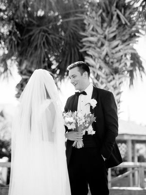 A black and white wedding photograph shows a couple sharing a tender moment outdoors among tropical foliage.