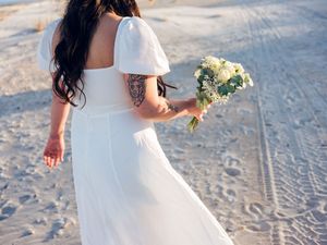 Close-up view of a bride's wedding dress swaying in the wind as she walks along a sandy beach with yellow flowers.