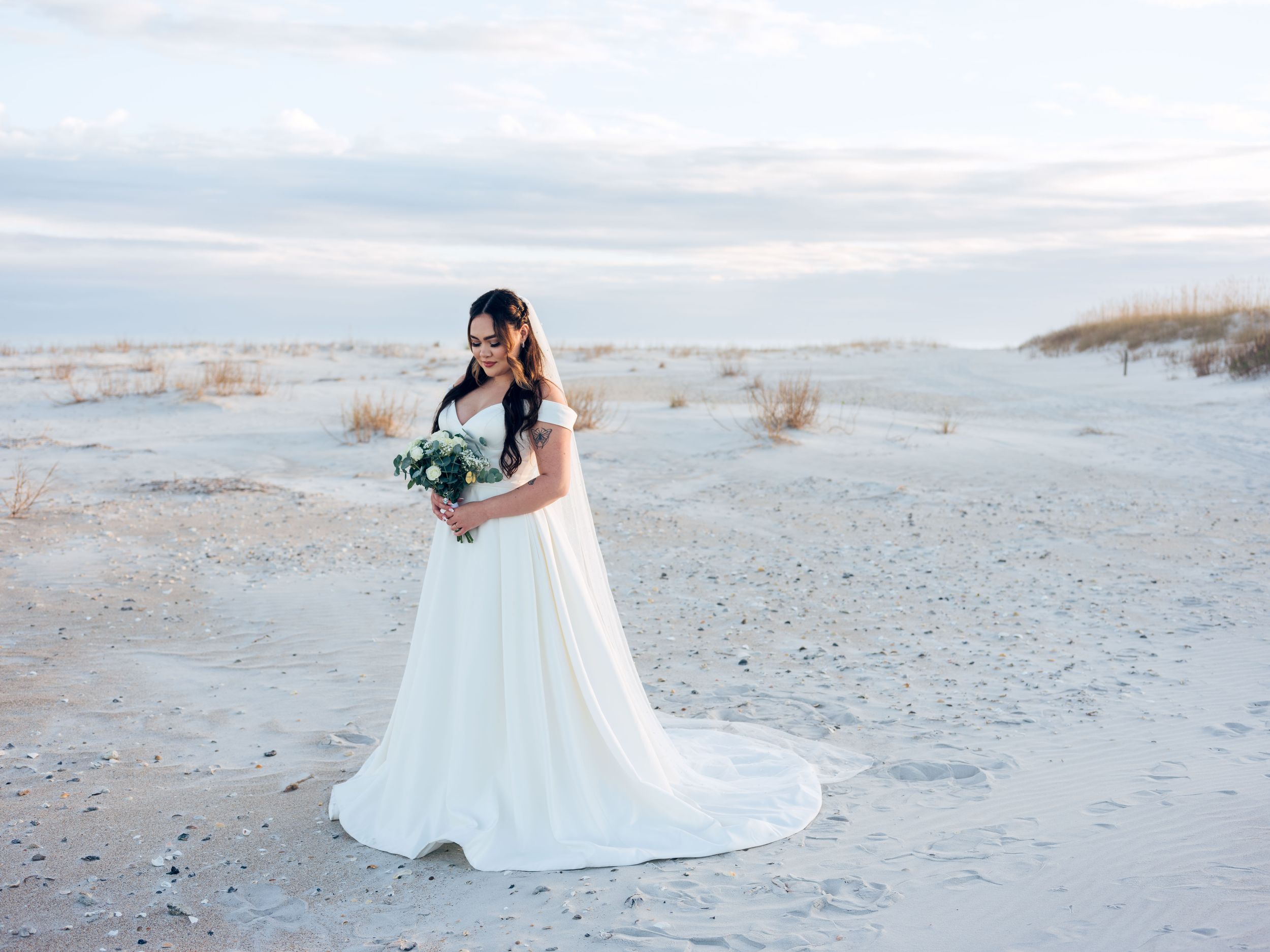 A couple in wedding attire poses together on a white sandy beach at sunset.