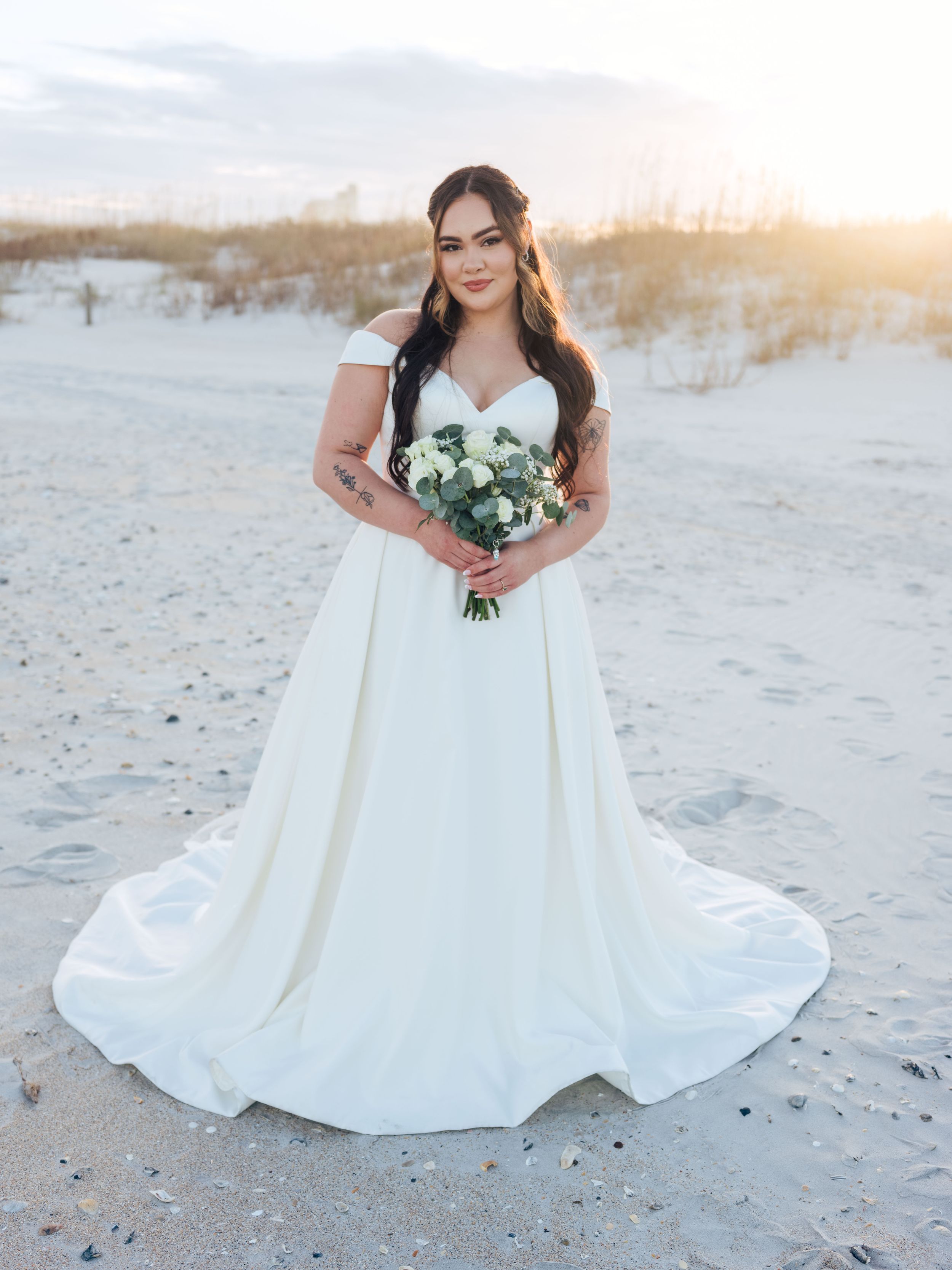 A bride in an elegant white ball gown poses gracefully on a sandy beach during golden hour sunset.