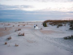 Wedding couple standing on white sand dunes at sunset with beach grass and cloudy sky along coastal shoreline.
