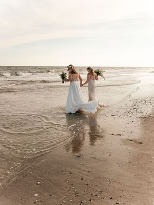 Couple in wedding attire sitting at the edge of ocean waves on sandy beach in romantic beach photoshoot.