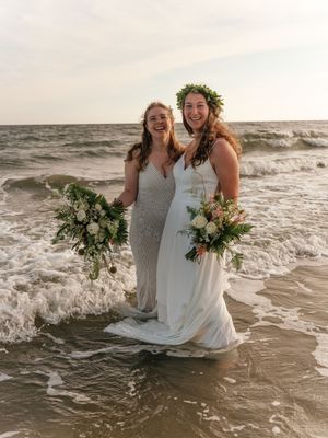 Two people in formal attire share a playful moment getting splashed by ocean waves at sunset on a sandy beach.