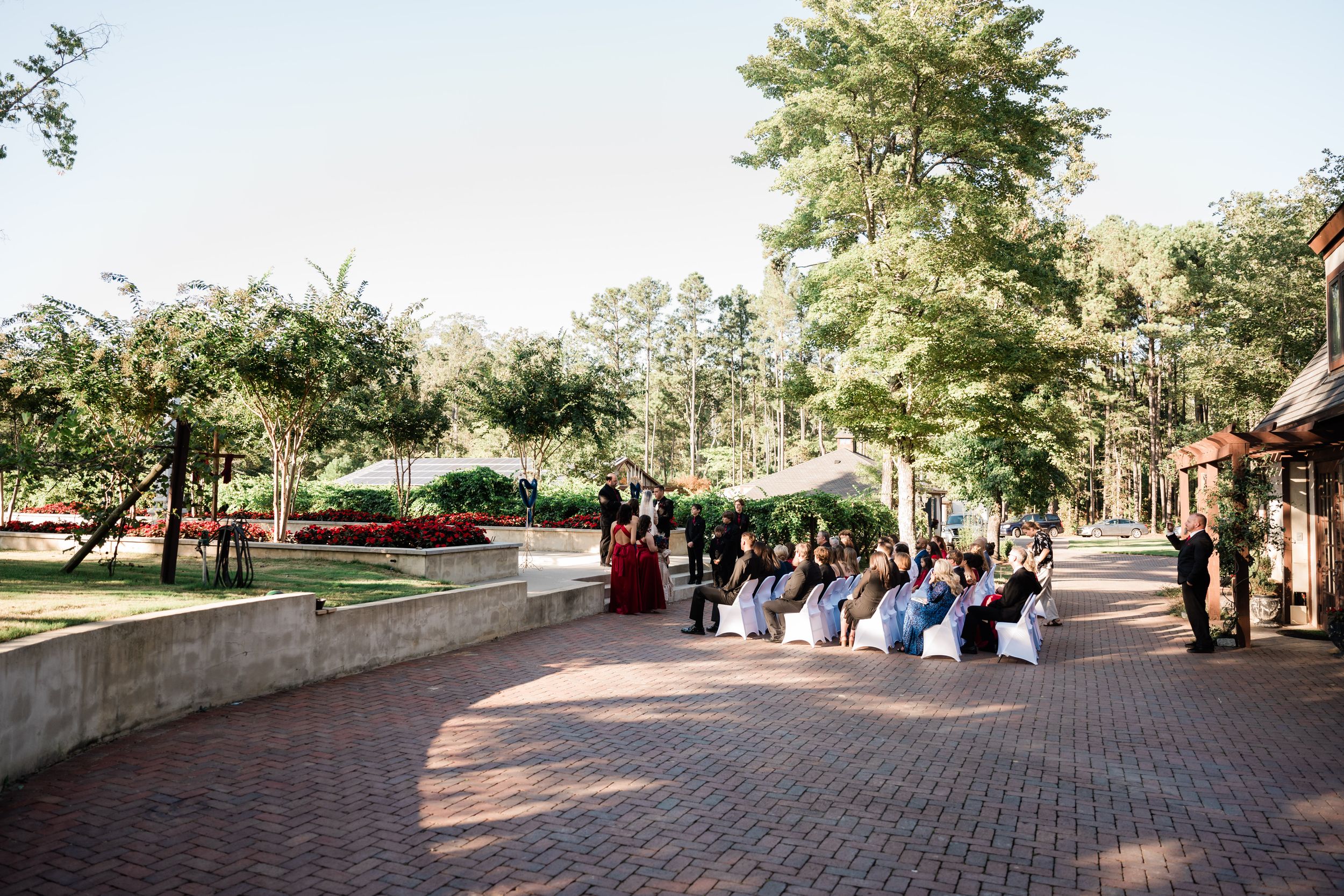 Wedding guests seated on brick walkway surrounded by lush greenery during outdoor ceremony.