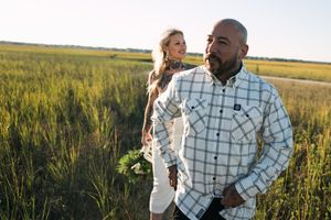 A couple stands together in a golden grass field at sunset as a breeze blows through the tall meadow.