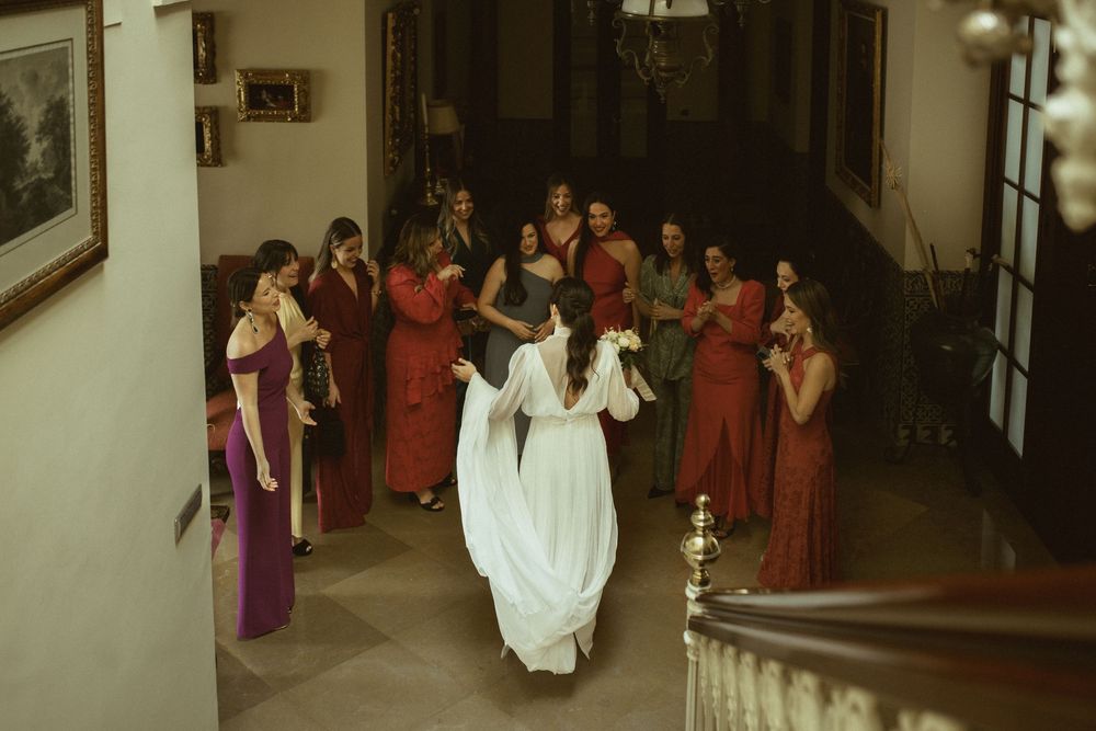 A bride in a white gown walks down a staircase surrounded by bridesmaids in burgundy dresses at an indoor venue.